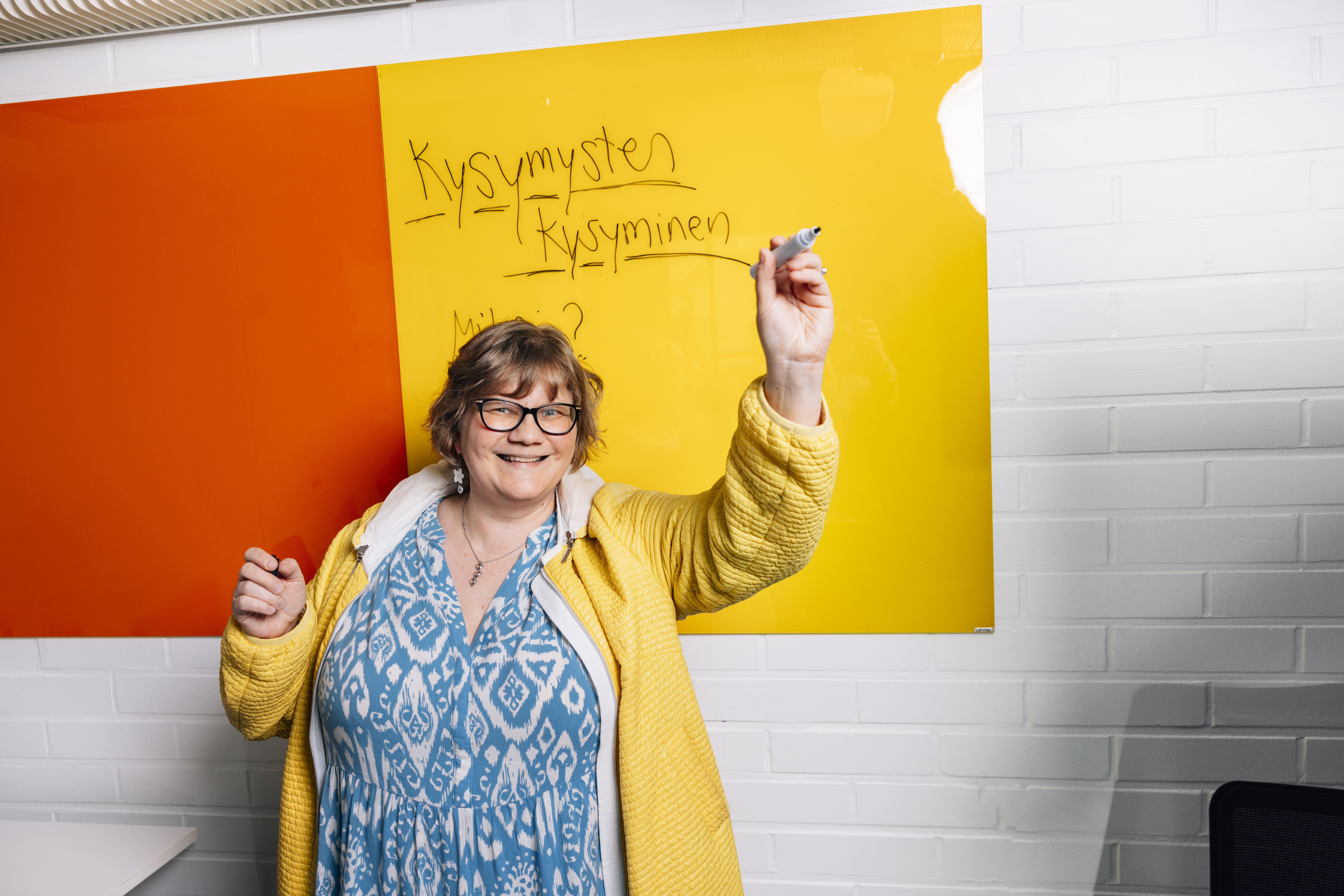 Nainen seisoo iloisesti taulun edessä pidellen kynää kädessään. Taustalla on oranssi ja keltainen taulu, johon on kirjoitettu tussilla tekstiä.