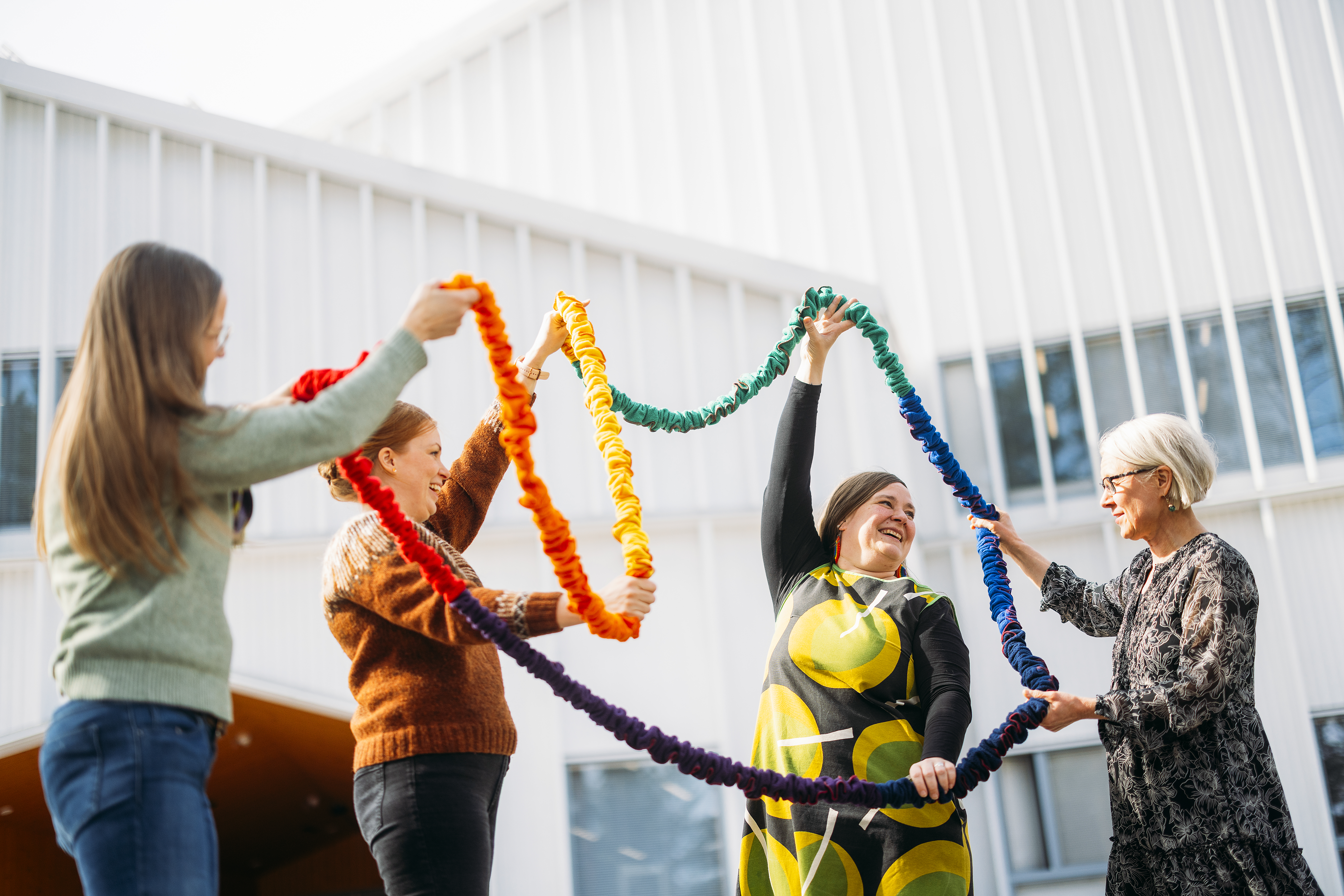 Neljä naista seisoo ulkona talon edustalla ja he pitelevät kädessään värikästä narua.
