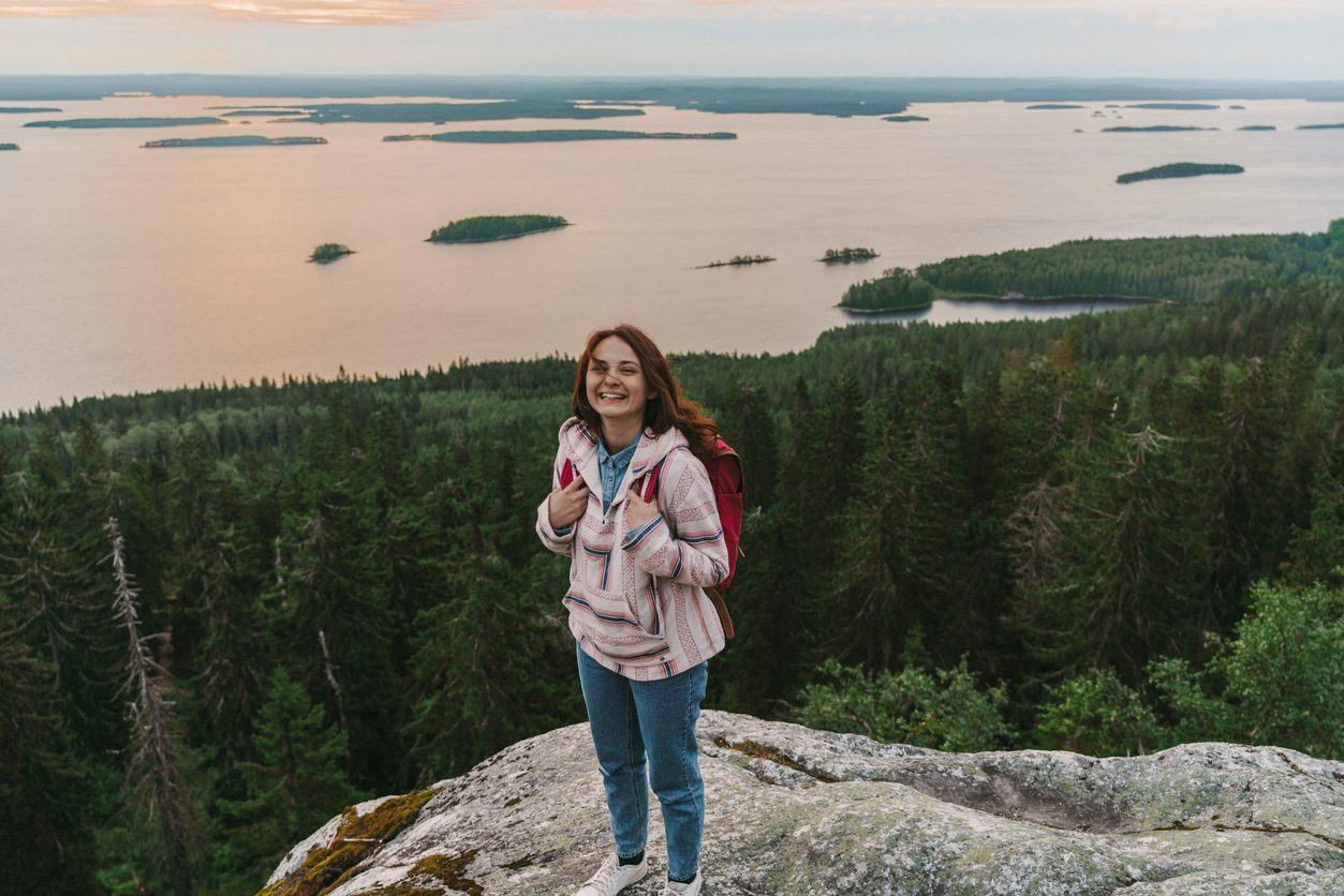 Nainen seisoo kallion laella ja katsoo laajaa metsä- ja järvimaisemaa.