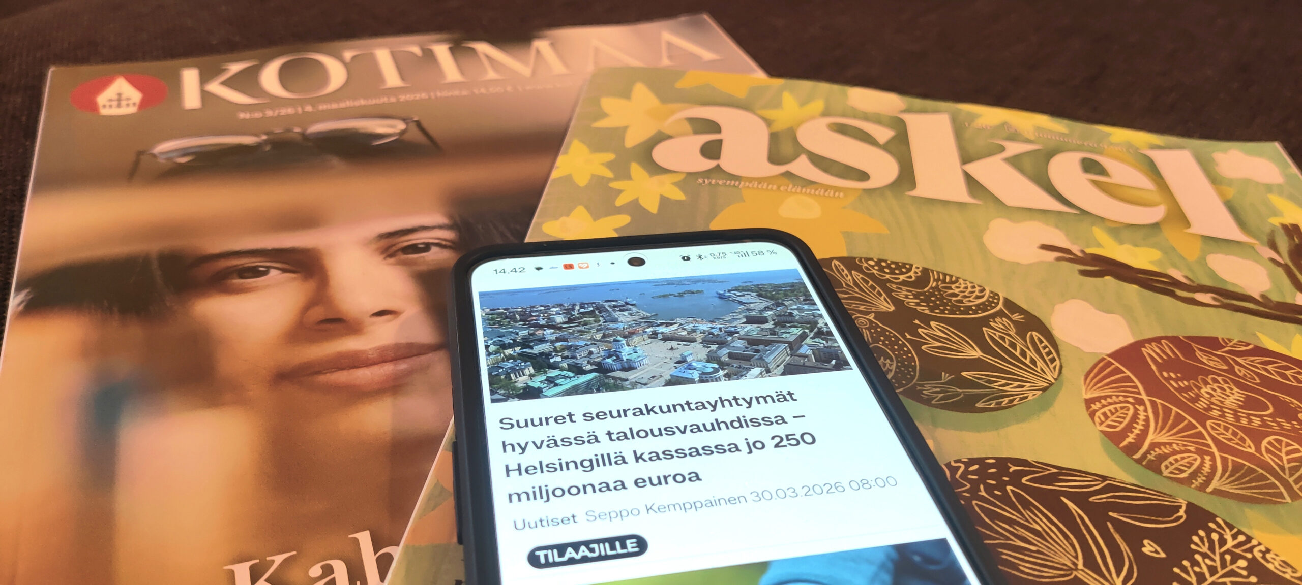 Pöydällä Kotimaa ja Askel sekä puhelin, jonka ruudulla on auki Kotimaa.fi-verkkomedian uutinen.