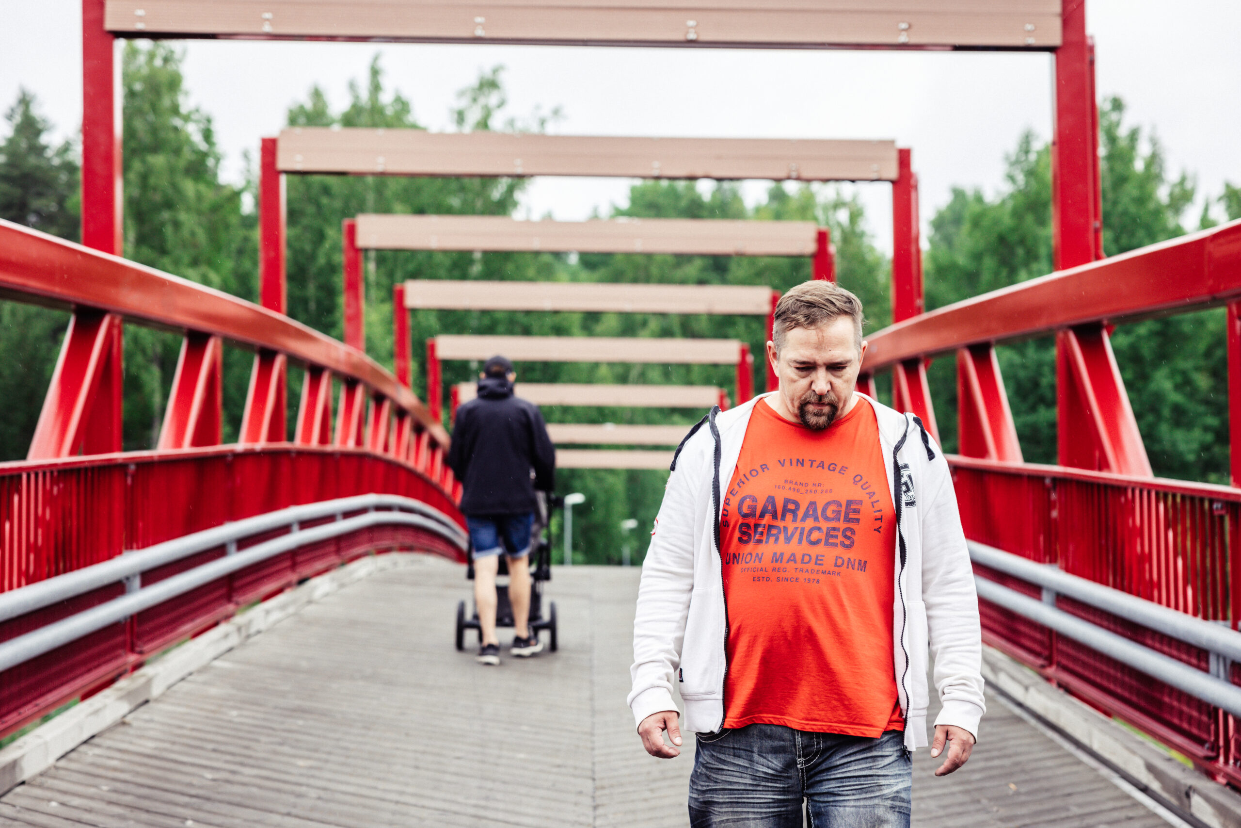Mies kävelee punaisella sillalla, taustalla toinen henkilö lastenrattaiden kanssa.
