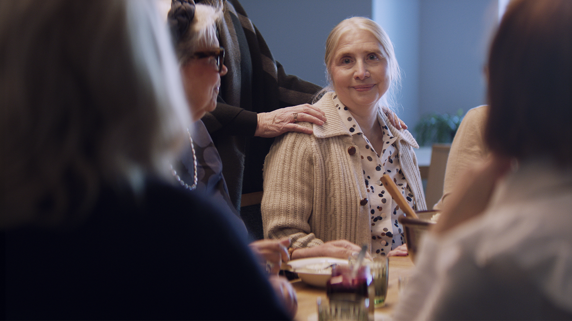 Ikääntyvä nainen on istunut pöytään, jossa on seuraa ympärillä. Yksi heistä on laskenut kädet vanhuksen hartioille.