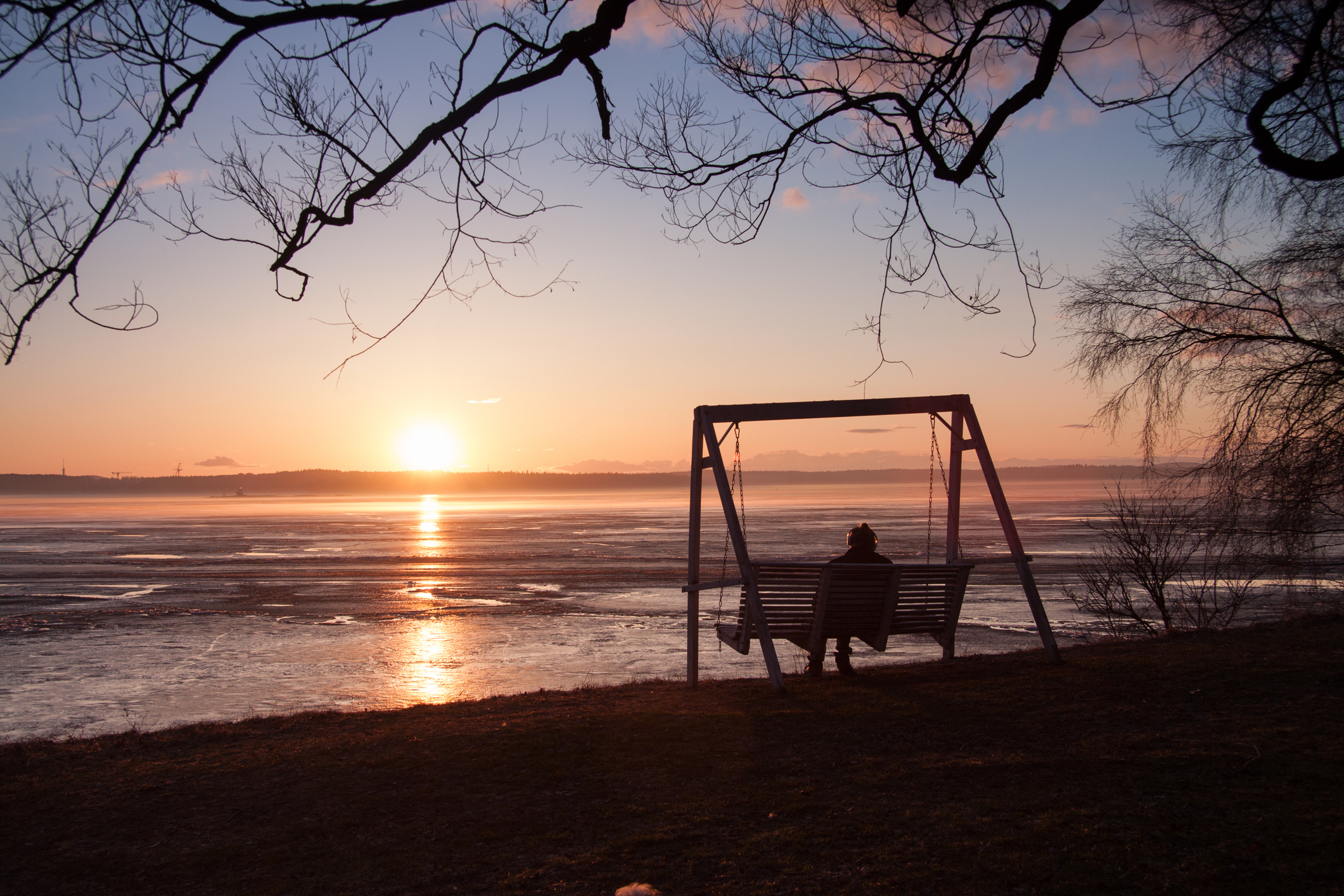 Nainen keinuu penkkikeinussa ja katsoo auringonlaskua jäätyneen järven yllä.